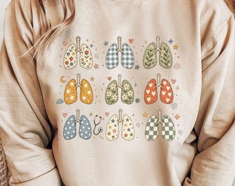 May include: A sand-colored crewneck sweatshirt featuring a graphic print of nine pairs of human lungs in various patterns, including floral, plaid, heart, and star designs, interspersed with small icons like a stethoscope, moon, and flowers.