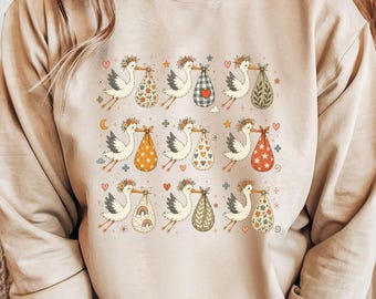 May include: A sand-colored crewneck sweatshirt featuring a repeating graphic print of nine storks, each wearing a floral crown and carrying a patterned bundle. The bundles display various designs including hearts, stars, flowers, and rainbows.