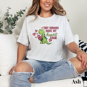 May include: A gray t-shirt with a green dinosaur holding a pink heart with the words "NICU" on it. The dinosaur is surrounded by pink hearts and the text "Tiny humans make my heart RAWr".