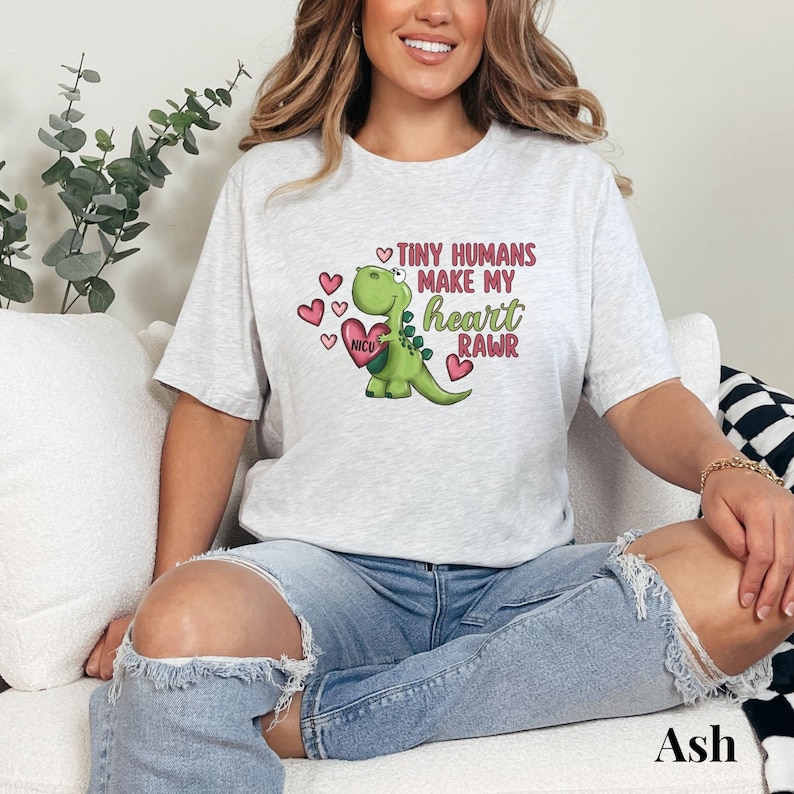 May include: A gray t-shirt with a green dinosaur holding a pink heart with the words "NICU" on it. The dinosaur is surrounded by pink hearts and the text "Tiny humans make my heart RAWr".