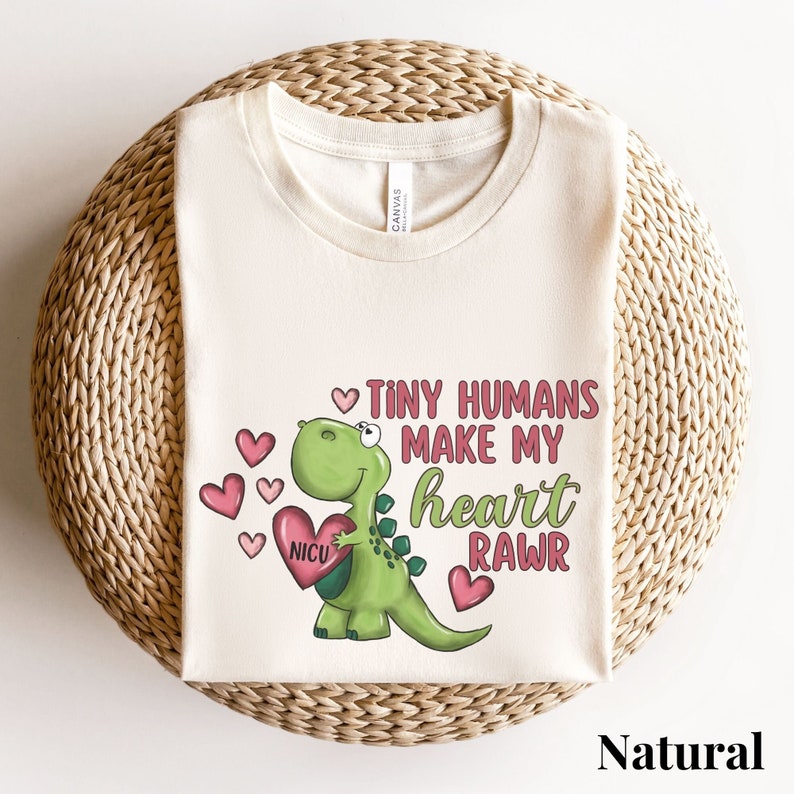 May include: A light beige t-shirt with a green dinosaur holding a pink heart with the word "NICU" on it. The dinosaur is surrounded by pink hearts. The text on the shirt reads "Tiny humans make my heart rawr".