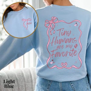 May include: Light blue sweatshirt with a pink ribbon and the text "Tiny Humans are my Favorite" in a pink frame.