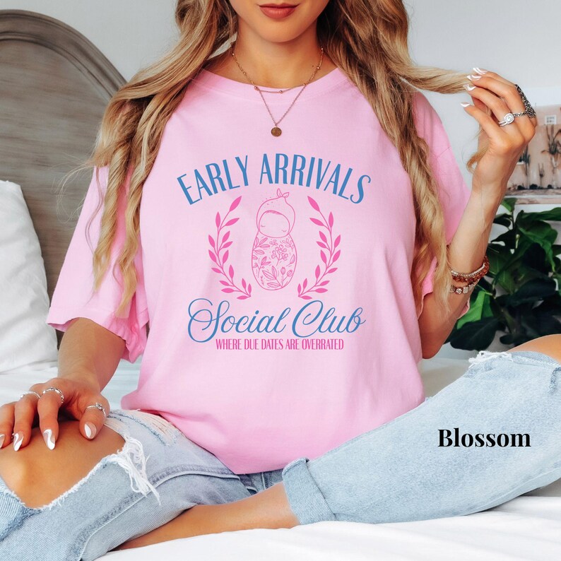 Op de afbeelding: Roze T-shirt met de tekst "Early Arrivals Social Club Where Due Dates Are Overrated" in blauwe en roze letters. Een kleine illustratie van een pop bevindt zich in het midden van de tekst.