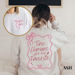 May include: A gray sweatshirt with a pink bow on the front and the text "Tiny Humans are my Favorite" in a pink frame on the back.