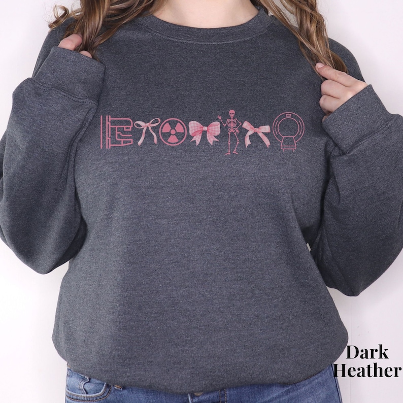 May include: Dark heather grey sweatshirt with a pink graphic of medical symbols including a stethoscope, a radiation symbol, a bow, a skeleton, and a CT scan.