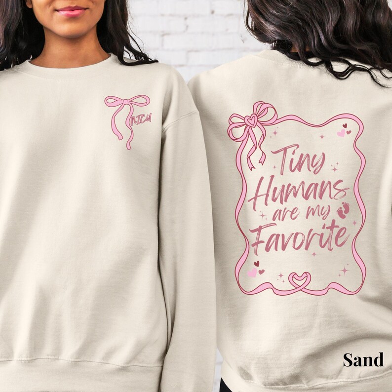 May include: A light beige sweatshirt with a pink bow on the left side and the text "Tiny Humans are my Favorite" with a pink bow and baby footprints on the right side.