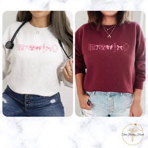May include: Two women wearing pink and white sweatshirts with a pink graphic of medical symbols. The sweatshirts are a light pink and a maroon color. The women are wearing blue jeans.