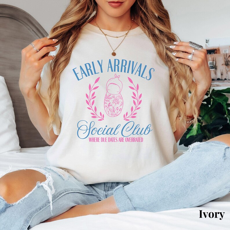 Op de afbeelding: Een wit T-shirt met een blauwe en roze afbeelding met de tekst "Early Arrivals Social Club Where Due Dates Are Overrated".