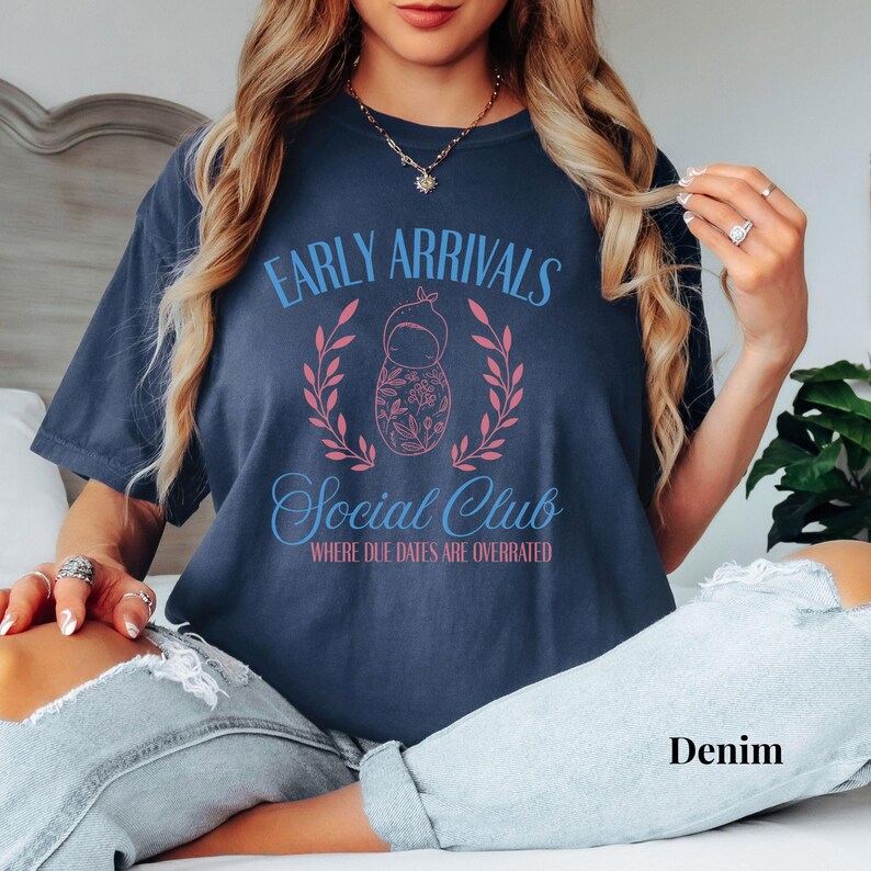 Op de afbeelding: Een marineblauw T-shirt met de tekst "Early Arrivals Social Club Where Due Dates Are Overrated" in roze en blauwe letters. Een roze en blauwe babypop-illustratie bevindt zich in het midden van de tekst.
