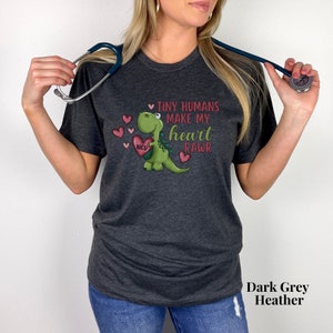 May include: A dark grey heather t-shirt with a green dinosaur holding a red heart with the text "NICU" on it. The dinosaur is surrounded by pink hearts and the text "Tiny humans make my heart RAWr".
