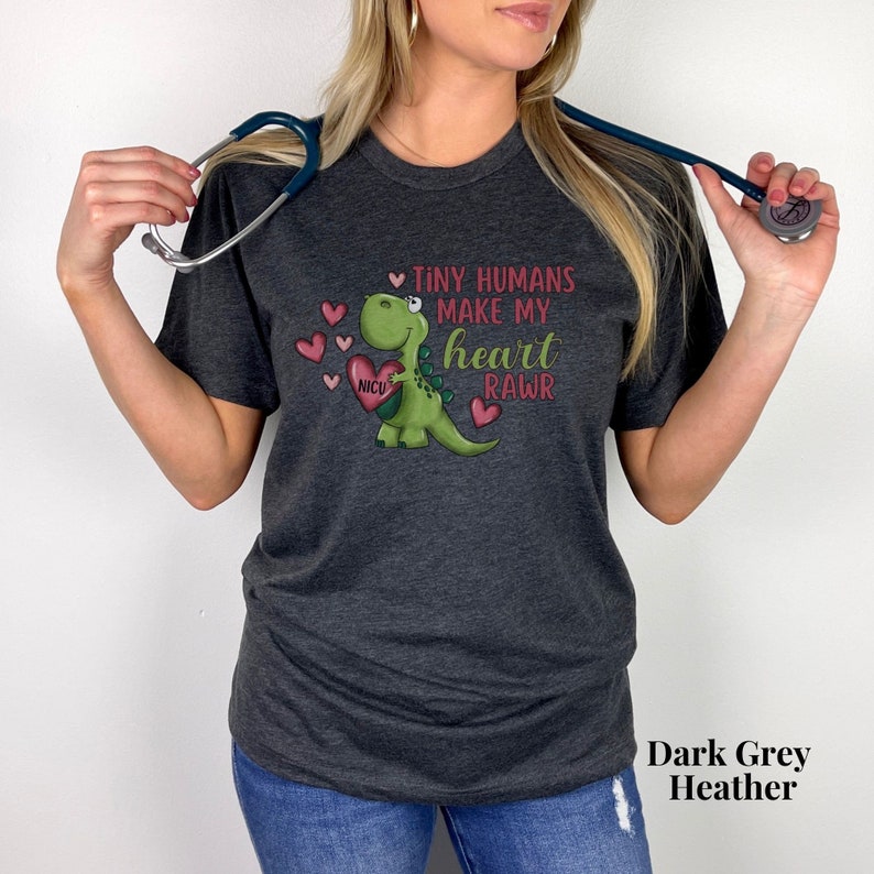 May include: A dark grey heather t-shirt with a green dinosaur holding a red heart with the text "NICU" on it. The dinosaur is surrounded by pink hearts and the text "Tiny humans make my heart RAWr".