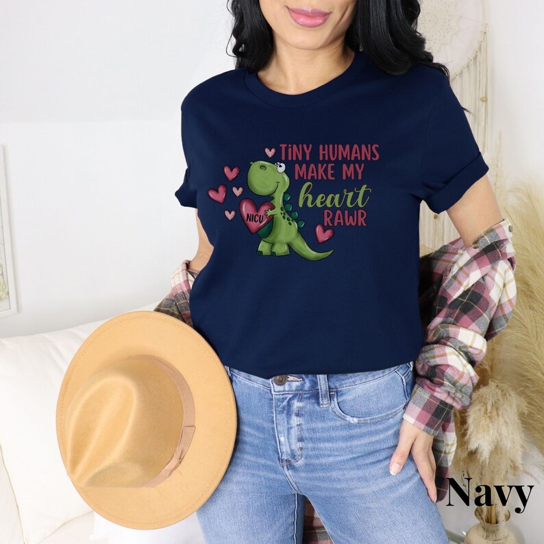 May include: Navy blue t-shirt with a green dinosaur holding a red heart with the text "NICU" on it. The dinosaur is surrounded by red hearts and the text "Tiny humans make my heart rawr".