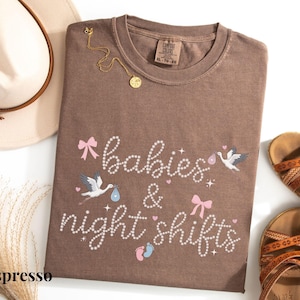K&ouml;nnte beinhalten: Espressofarbenes T-Shirt mit dem Schriftzug "babies & night shifts" in Schreibschrift, verziert mit rosa Schleifen, Storch-Illustrationen und Babyf&uuml;&szlig;en. Eine goldene Halskette und ein beiger Hut mit braunem Band sind ebenfalls im Bild, zusammen mit braunen Sandalen.