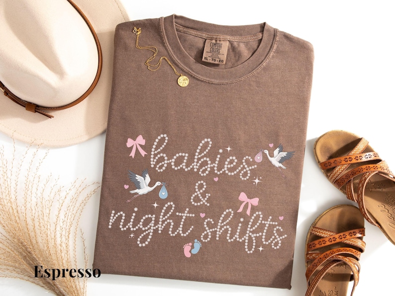 K&ouml;nnte beinhalten: Espressofarbenes T-Shirt mit dem Schriftzug "babies & night shifts" in Schreibschrift, verziert mit rosa Schleifen, Storch-Illustrationen und Babyf&uuml;&szlig;en. Eine goldene Halskette und ein beiger Hut mit braunem Band sind ebenfalls im Bild, zusammen mit braunen Sandalen.