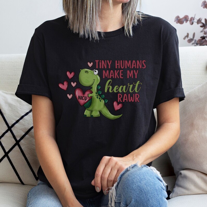 May include: Black t-shirt with a green dinosaur holding a red heart with the word "NICU" on it. The text on the shirt reads "Tiny humans make my heart rawr".