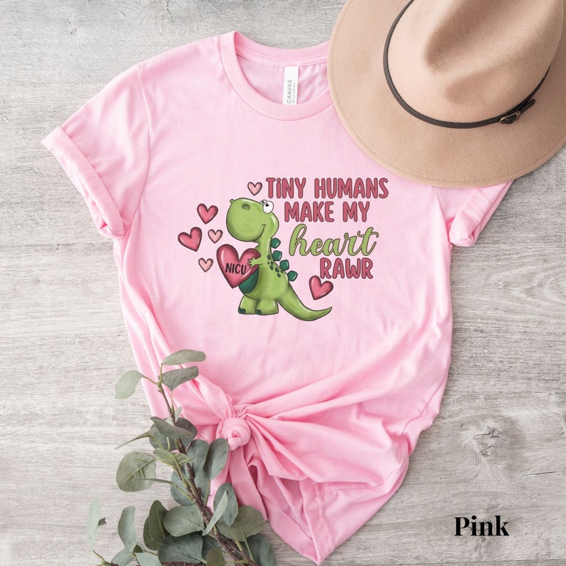 May include: Pink t-shirt with a green dinosaur holding a heart that says "NICU". The text on the shirt reads "Tiny humans make my heart rawr".