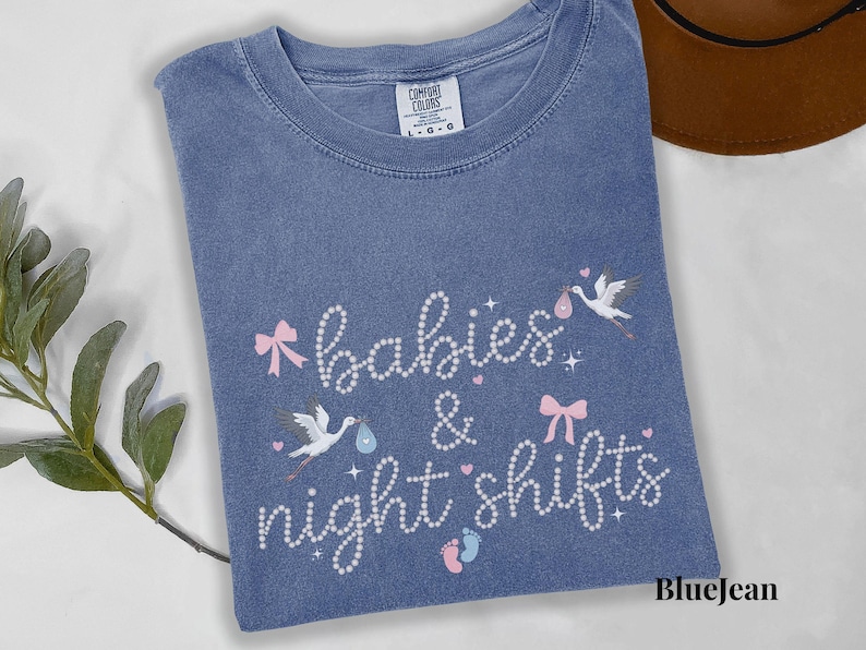 K&ouml;nnte beinhalten: Blaues Jeans-T-Shirt mit den Worten "babies & night shifts" in Schreibschrift. Das Design umfasst rosa Schleifen, St&ouml;rche, die Babys tragen, und Babyf&uuml;&szlig;e. Das Shirt ist aus einem weichen Material.