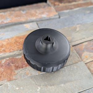 May include: A black, circular object with a ridged edge and a central, raised section featuring a triangular opening. The object appears to be made of a solid material, possibly plastic or metal, and is designed for a specific mechanical purpose.