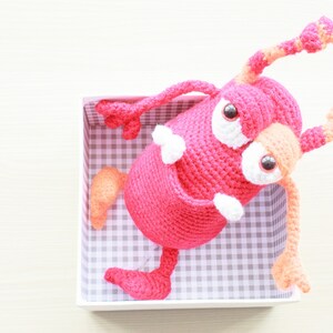 Könnte beinhalten: Ein rosa gehäkeltes Monster-Spielzeug mit orangefarbenen und weißen Akzenten. Das Spielzeug hat große Augen und sitzt in einer weißen Schachtel mit einem karierten Muster.