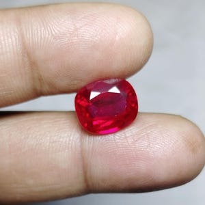 May include: A vibrant, cushion-cut ruby gemstone. The gem is a deep, saturated red color, with facets that catch the light. The ruby is held between two fingers, showcasing its size and brilliance. This gemstone is suitable for jewelry.