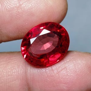 May include: A faceted, oval-shaped, deep red gemstone. The gem is held between fingers, showcasing its polished surface and internal reflections. The stone's color is a vibrant, saturated red, with a high degree of clarity.