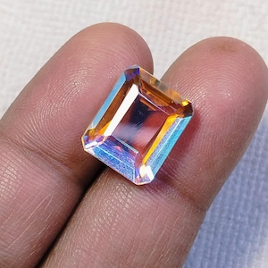 May include: An emerald-cut gemstone with a vibrant display of colors, including orange, blue, and yellow. The gem is held in a hand, showcasing its facets and reflective qualities. The gemstone's rectangular shape and polished surface are clearly visible.