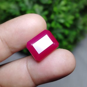 May include: A vibrant, rectangular-cut gemstone with a deep magenta-pink hue and faceted surfaces that reflect light. The stone features sharp, clean edges and a polished finish, showcasing a uniform, saturated colour throughout its entire structure.
