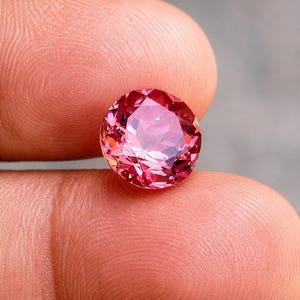 May include: A round, faceted gemstone with a vibrant pink hue. The stone features a brilliant cut with numerous triangular and kite-shaped facets that reflect light, creating a sparkling appearance with deep magenta and lighter rose-colored tones.