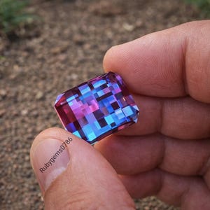 May include: A rectangular gemstone with multiple facets, showcasing a mosaic of blue, purple, and pink hues. The gem is held between fingers, with the text "rubygems0786" visible on a finger.
