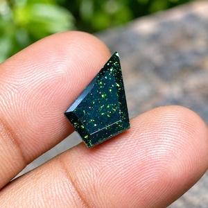 May include: A faceted shield-shaped gemstone with a deep dark green base color and vibrant, sparkling lime green flecks distributed throughout the surface. The stone features sharp, geometric edges and a polished, reflective finish that catches light.