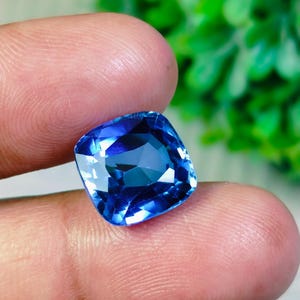 May include: A cushion-cut gemstone with a deep blue color and a faceted surface that reflects light. The stone has a square shape with rounded corners and a transparent appearance, showcasing internal clarity and a vibrant, saturated blue hue.