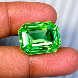 Puede incluir: Una gema de corte esmeralda con un color verde lima vibrante y saturado. La piedra presenta facetas geométricas precisas que crean reflejos de luz nítidos y una profunda claridad interna, mostrando una forma rectangular con esquinas biseladas.