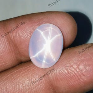 Op de afbeelding: Een ovaalvormige, doorschijnende, lichtroze stersaffier edelsteen met een duidelijk zesstralig stereffect, bekend als asterisme, dat verschijnt als een heldere witte lichtreflectie in het midden van het gladde, gepolijste cabochon-oppervlak.
