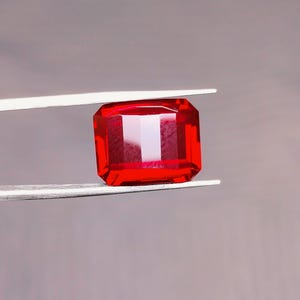 May include: A vivid, rectangular-cut, red gemstone is held by silver tweezers against a grey background. The gem's facets reflect light, showcasing its clarity and rich colour. This gemstone is likely for jewellery making.