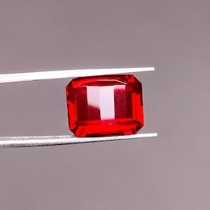 Puede incluir: Una gema de color rojo vibrante con corte esmeralda, sostenida por unas pinzas de metal plateado. La gema presenta una forma rectangular con bordes facetados que reflejan la luz, creando una apariencia brillante y clara sobre un fondo neutro.