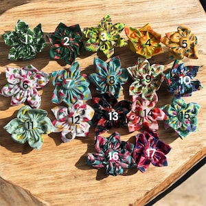 Può includere: Una collezione di diciassette abbellimenti floreali in tessuto in vari colori e motivi. Ogni fiore ha un design unico, inclusi motivi floreali e geometrici. I fiori sono disposti su una superficie di legno, mostrando i loro diversi stili.