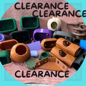 Puede incluir: Una colección de diversos hábitats y accesorios para animales pequeños en una variedad de colores y formas. Los artículos incluyen recintos verdes, morados, marrones y grises, un escondite en forma de calabaza y un plato en forma de corazón. La palabra "CLEARANCE" está escrita en negro.