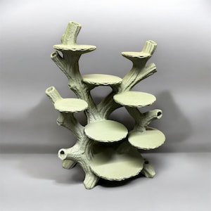 May include: A light green display stand shaped like a tree with multiple branches and seven circular platforms at varying heights. The structure features a textured, bark-like surface throughout, providing a tiered arrangement for items.
