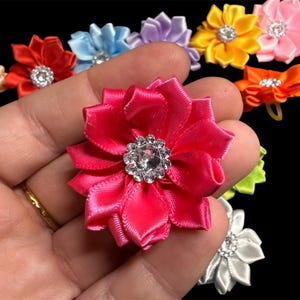 May include: A selection of colourful satin flower embellishments. The flowers are in various colours, including pink, red, blue, yellow, and orange. Each flower has a sparkling rhinestone centre. The background is black.