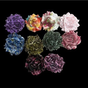 May include: A selection of ten fabric flower embellishments in diverse colours and patterns. The flowers feature shades of purple, orange, white, pink, blue, green, and burgundy. Some have polka dots or floral prints, while others have textured designs.