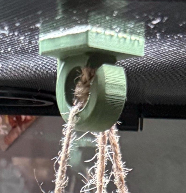 May include: A green, 3D-printed plant hanger with a circular opening and a textured design. The hanger is attached to a woven rope, suitable for suspending a plant pot. The background is blurred, suggesting an indoor setting.