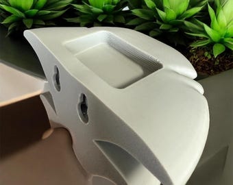 May include: A light gray, abstract-shaped wall planter with a rectangular basin and two circular holes for mounting. The planter is positioned in front of several green plants.