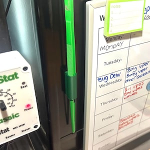 Może przedstawiać: Jasnozielona pęseta do karmienia gadów z uchwytem przymocowanym do ciemnej powierzchni. Na pęsecie wydrukowano napis "International Rescue". Biały kalendarz z odręcznymi notatkami znajduje się w tle.