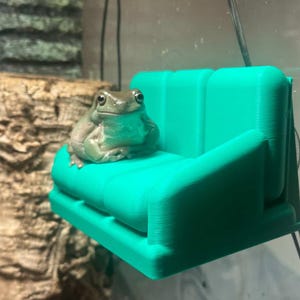 以下が含まれることがあります： 小さなターコイズ色の3Dプリントのソファに、緑色の木のカエルが座っています。カエルは薄い緑色で、目は濃い色をしています。ソファは、平らな座面と背もたれを備えたシンプルなデザインです。