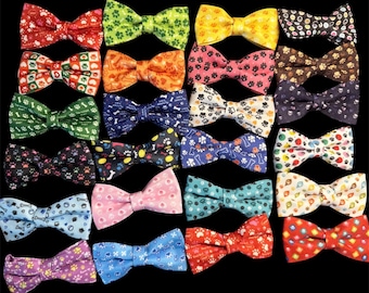 May include: A collection of twenty-five bow ties arranged in a grid, each featuring various colourful patterns including paw prints, bones, and geometric shapes in red, green, yellow, blue, pink, black, and white on a solid black background.