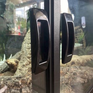 Könnte beinhalten: Schwarze, gebogene Griffe an einer Glastür eines Terrariums. Die Griffe sind rechteckig mit einer vertieften Griffmulde. Der Hintergrund zeigt einen natürlichen Lebensraum mit Holz und Pflanzen.