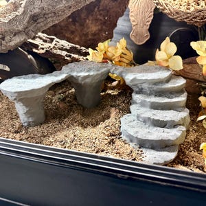 Op de afbeelding: Een terrariumopstelling met een lichtgrijze rotsbrug en trappen, omringd door zand en kunstmatige gele bloemen. Een stuk drijfhout en een decoratieve zebrahoofd zijn ook zichtbaar.