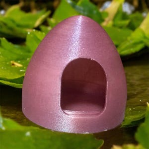 Puede incluir: Un pequeño objeto en forma de cúpula de color malva con una abertura redondeada. El objeto tiene una superficie texturizada y se encuentra sobre un fondo de hojas verdes. El diseño se asemeja a un refugio o cueva en miniatura.