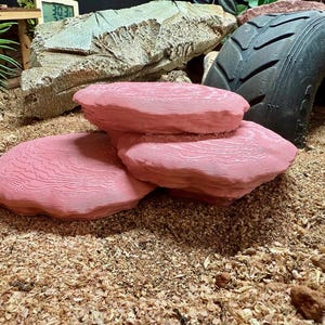 May include: Three pink artificial rocks with a textured surface, stacked to resemble natural formations. They sit on wood shavings, possibly for a terrarium or display. The rocks are a vibrant pink colour.