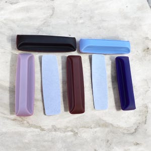 May include: An assortment of rectangular, colorful objects in various shades, including black, blue, purple, and burgundy. Some have a smooth, glossy finish, while others appear textured. The objects are arranged on a light-colored surface.